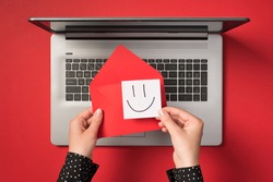 First person top view photo of hands holding red vivid envelope and white sticker note paper with drawn smiling face over open grey laptop on isolated red background