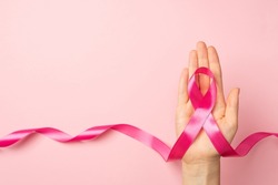 First person top view photo of female hand holding pink ribbon in palm symbol of breast cancer awareness on isolated pastel pink background with copyspace