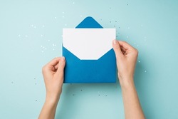 First person top view photo of hands holding open blue envelope with white card over sequins on isolated pastel blue background with empty space
