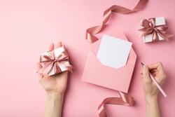 First person top view photo of st valentine's day decor female hands holding pen pink envelope with letter small giftbox and pink silk curly ribbon on isolated pastel pink background with empty space