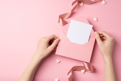 First person top view photo of valentine's day decor young woman's hands holding open pink envelope with paper card hearts and pink silk curly ribbon on isolated pastel pink background with copyspace
