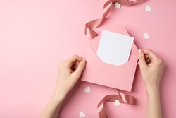 First person top view photo of valentine's day decor girl's hands holding pink envelope with letter heart shaped confetti and pink satin curly ribbon on isolated pastel pink background with copyspace