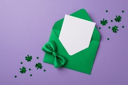 Top view photo of saint patricks day decor clover shaped confetti green envelope with paper card and green bow tie on isolated pastel lilac background with empty space