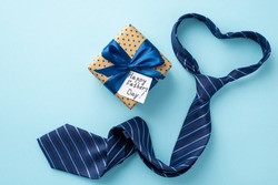 Father's Day concept. Top view photo of heart shaped blue necktie polka dot giftbox with satin ribbon bow and postcard on isolated pastel blue background