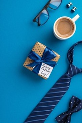 Father's Day concept. Top view vertical photo of polka dot gift box with ribbon bow and postcard blue necktie bow tie glasses cup of hot drinking and cufflinks on isolated blue background