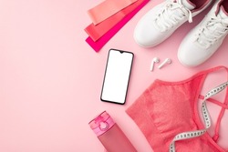 Fitness concept. Top view photo of white sneakers pink sports bra resistance bands tape measure bottle of water smartphone and earbuds on isolated pastel pink background with copyspace