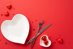 Valentine's Day concept. Top view photo of heart shaped plates cutlery knife fork chocolate candies and confetti on isolated red background with empty space