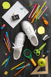 Education-themed display. High-angle vertical shot capturing a pair of white sneakers, copybook, apple snack and a wide range of learning materials on a chalkboard surface background