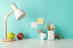 Empower students with an organized study zone through this side-view picture featuring white desk with lively school supplies in penholder and lamp on a blue backdrop, suitable for text or ad usage