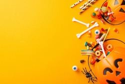 Enjoyable children's trick or treating tradition. Top-down image of a pumpkin basket with candies and Halloween decorations on orange isolated backdrop, providing copy-space for text or advertising