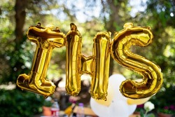 Balloons in the shape of the letters IHS, referring to the sacrament of First Holy Communion. In the background, an outdoor party for children. IHS 