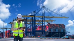 Engineer wearing uniform inspection and see detail on tablet with logistics container dock cargo yard with working crane bridge in shipyard with transport logistic import export background.
