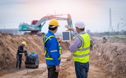 Construction engineer wear safety uniform under inspection and survey workplace by tablet with excavation truck digging and worker construction road background.