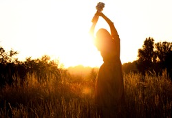 Silhouette of a woman with long natural hair in a long mustard-colored cotton dress picks wild flowers against the background of the sunset. Natural beauty, privacy in nature, women's day.