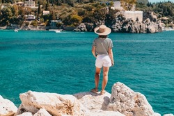 Back view of woman in striped cruise outfit, straw hat. Lady standing on stone beach. Teal sea background. South Europe Riviera, travel destination, luxury vacation concept.