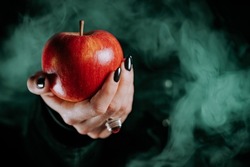 Woman as witch offers red apple as symbol of temptation, poison. Fairy tale, white snow wizard concept. Spooky halloween, cosplay. Smoke, haze background.