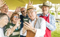 A group of retired friends enjoys using technology during an outdoor barbecue. The seniors enjoy using technology, taking selfies and making video calls.