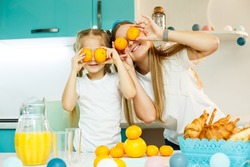 A young mother and daughter in the kitchen at the table play with fresh citrus fruits, tangerines instead of eyes on their faces. Family relationship with a child