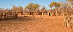 Wide angle view of a small Himba village in rural Namibia, with traditional round mud houses, thatched roofs and wooden goat pens in a sandy, arid landscape.