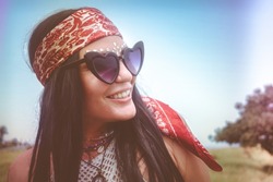 Matte, retro style, close-up portrait of a cool, artsy, bohemian woman with long straight black hair, vintage bandanna, beaded jewelry, face glitter and large heart shaped sunglasses.