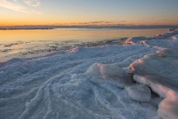 Snow and ice on frozen river coast near sea water waves and cold winter evening sunset sky clouds reflection