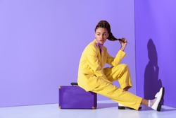 african american girl posing in yellow suit sitting on purple suitcase, on trendy ultra violet background