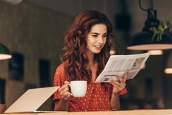 portrait of woman with cup of coffee reading newspaper at table with laptop in cafe