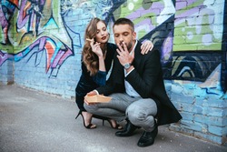 couple in fashionable outfit with french fries sitting near wall with graffiti on street