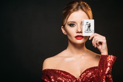 attractive girl in red shiny dress covering eye with joker card isolated on black
