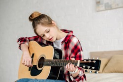 Concentrated teenager in checkered shirt playing acoustic guitar at home