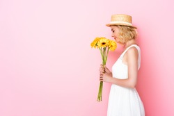 side view of woman in straw hat and white dress smelling yellow flowers on pink