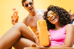 young interracial couple with orange juice and cocktail looking at camera isolated on yellow
