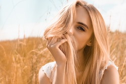 young blonde woman obscuring face with hair while looking at camera against blue sky in field