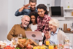 Selective focus of multicultural family having video call on laptop near food during thanksgiving