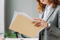 Cropped view of businesswoman leafing through paper sheets in folder with blurred workplace on background