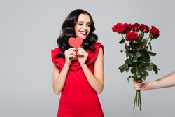 man giving roses to happy woman in red dress holding heart-shaped card isolated on grey