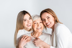 three generation of positive women smiling while looking at camera and hugging isolated on grey