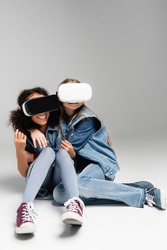frightened and thrilled multicultural kids in vr headsets embracing while sitting on grey