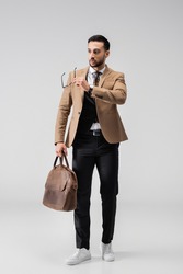 full length view of fashionable arabian man holding eyeglasses and leather bag on grey