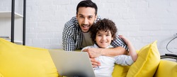 happy muslim man hugging smiling son sitting on sofa with laptop, banner