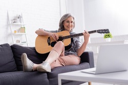 cheerful middle aged woman playing acoustic guitar on couch near laptop on coffee table
