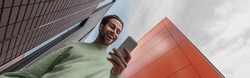 low angle view of amazed man in sweatshirt holding smartphone near buildings, banner