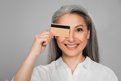 cheerful middle aged woman with grey hair obscuring eye with credit card isolated on grey
