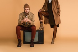 young man in autumn jacket and beanie sitting on vintage tv set near woman in stylish boots on beige background