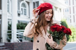 happy young woman in trench coat holding red roses and adjusting beret on urban street of europe