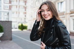 happy young woman in black turtleneck and leather jacket adjusting hair on urban street of europe