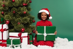 African american child in santa hat holding gift near christmas tree and decorative snow on green background