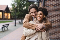 happy man embracing amazed african american girlfriend near building outdoors