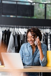 cheerful african american saleswoman talking on smartphone and using laptop in clothing boutique