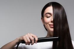 pleased brunette woman using hair straightener isolated on grey